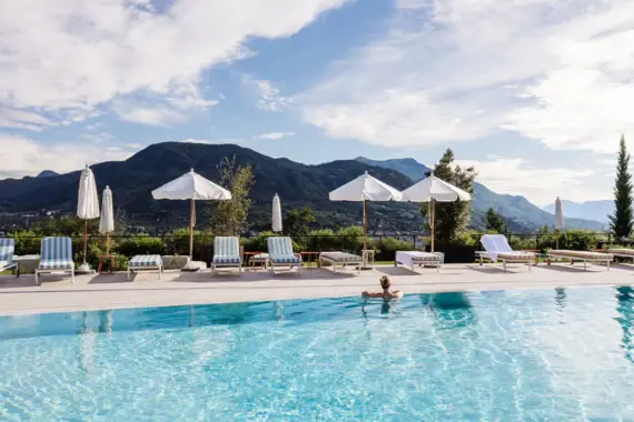 Persona appoggiata al bordo di una piscina con ombrelloni, sedie a sdraio a righe e montagne sullo sfondo.