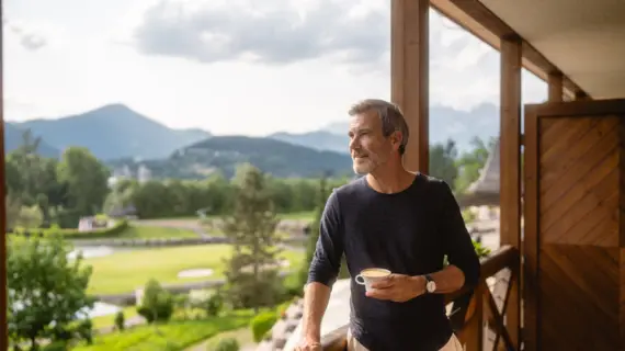 Un uomo è in piedi su un balcone, con una tazza di caffè in mano. Guarda in lontananza un paesaggio montano.