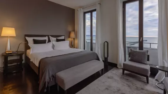 Un'elegante camera da letto con vista sul mare al tramonto. Al centro della stanza si trova un grande letto con biancheria grigia e cuscini bianchi e grigio scuro. Ai lati del letto si trovano comodini con lampade da tavolo che emettono una luce calda, accanto ai quali si trova una comoda poltrona.