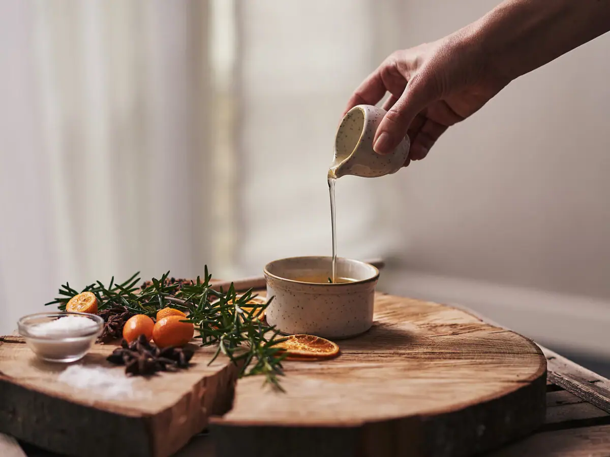 Eine Person gießt Massageöl in eine Schüssel, um die herum eineige Kräuter auf einem Holzbrett platziert sind..