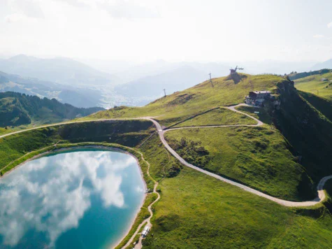 Lago situato su una collina con un edificio sulla cima, circondato da montagne e prati verdi.