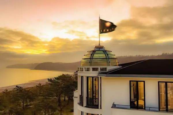 Una bandiera con la scritta "CERES" sventola al vento su una cupola di vetro di un edificio bianco. Sullo sfondo si vedono alberi, la spiaggia di Binz e il Mar Baltico al tramonto.