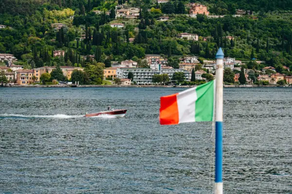 Flagge auf einem Mast vor einem Boot auf dem Wasser und grün bewachsenen Hügeln im Hintergrund..