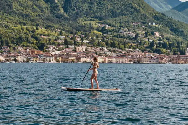 Eine Frau fährt mit einem Stand-up-Paddle Board auf dem Garadsee vpr der Kulisse einer Stadt vor grün bewachsenen Hängen an der Küste.