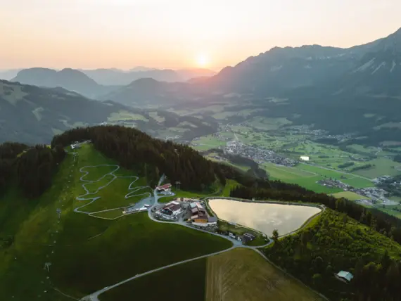 Una veduta aerea di montagne verdi e di una strada tortuosa lungo un lago. Sullo sfondo dell'immagine si vede una valle con un piccolo villaggio. 