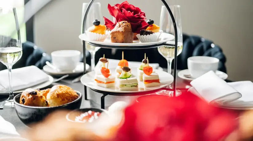 Un tavolo da pranzo con un'etagere di piccoli snack e pasticcini, accanto al quale sono disposti tazze da caffè, bicchieri da champagne e piatti. 