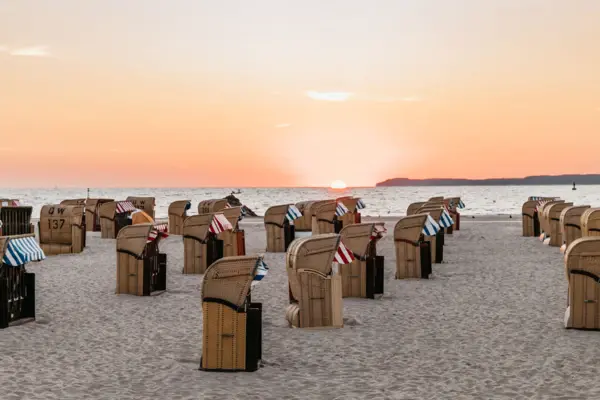 Una spiaggia con un mare dolcemente ondulato, piena di sedie a sdraio rosse e blu, mentre il sole sorge e bagna il cielo di un caldo colore arancione. 