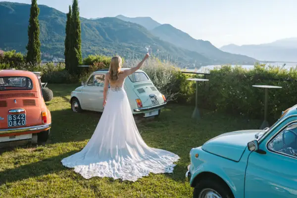 Eine Frau im Hochzeitskleid hält ein Glas Champagner vor einem Fiat 500 Auto.