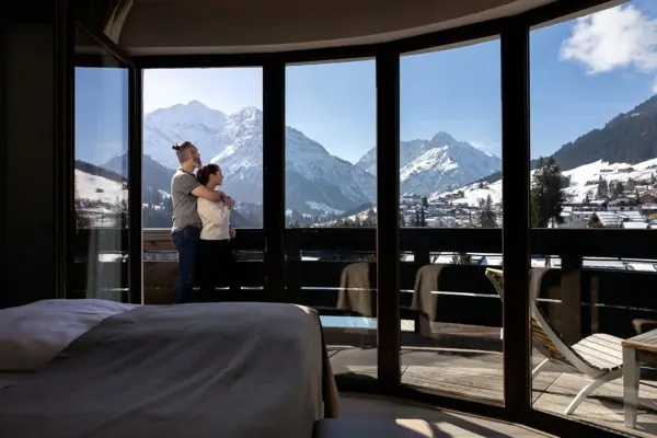 Camera d'albergo Una coppia è in piedi davanti a una grande finestra che si affaccia su un paesaggio montano innevato.