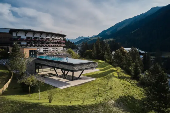 A-ROSA Ifen Hotel Kleinwalsertal Un edificio con una piscina al centro, circondato dalle montagne e dal cielo azzurro.