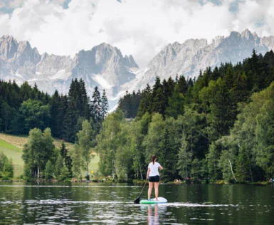 Donna su una tavola da paddle in un lago, circondata da montagne e alberi.