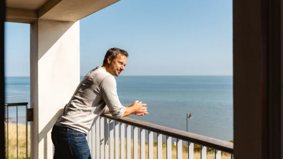 Uomo in jeans appoggiato alla ringhiera su un balcone che guarda il mare.