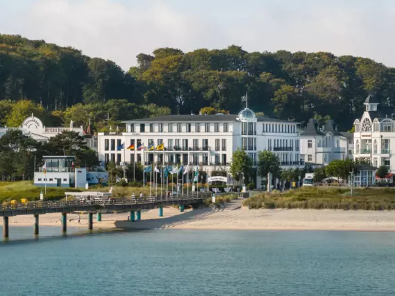 Un edificio bianco con finestre a cornice nera, una cupola di vetro e una bandiera sul tetto si erge davanti a una fitta foresta di faggi sulla costa di Binz. Il Mar Baltico è visibile in primo piano, così come un tratto della spiaggia di Binz e il molo di Binz, che si protende nel Mar Baltico.