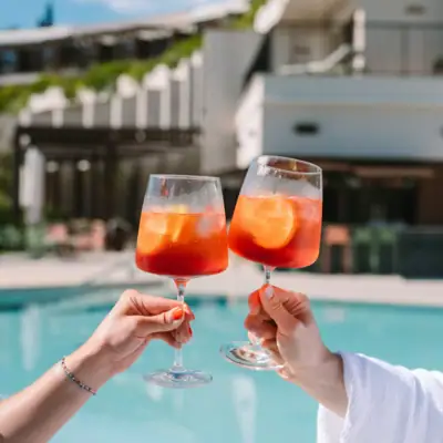 Mani che tengono bicchieri di vino con liquido arancione e ghiaccio.