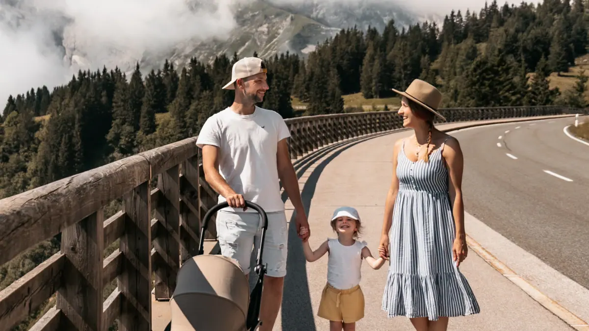 Una famiglia attraversa un ponte con un passeggino; sullo sfondo si vede una montagna.