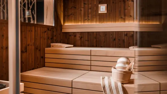 Una sauna con due panche in una luce accogliente. Sulla panca inferiore della sauna si trovano un secchio di legno per gli infusi e due teli da sauna a righe.