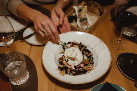 Persona che mangia un pasto su un piatto, con bicchiere di vino sul tavolo.