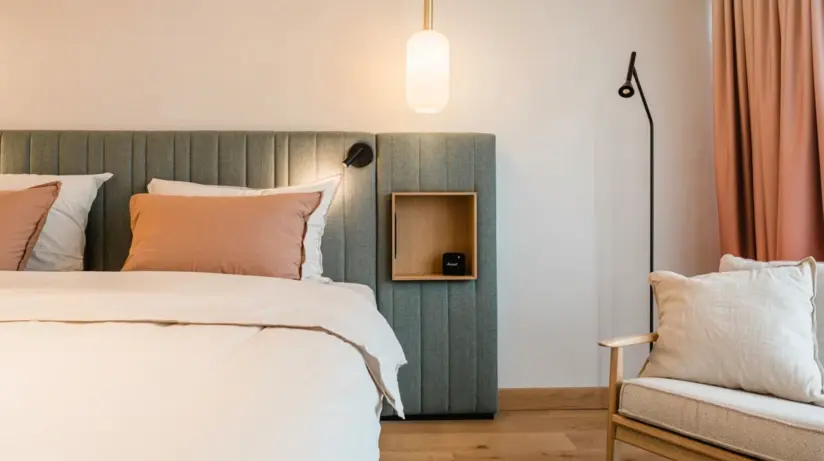 Una camera da letto dettagliata con un ampio letto matrimoniale, la cui testata è rivestita in tessuto e rifinita in un elegante grigio. Delicati cuscini rosa e beige completano la biancheria da letto bianca. Una lampada a sospensione minimalista fluttua sopra il comodino e si inserisce armoniosamente nell'ambiente moderno. Un'elegante lampada da terra nera e una comoda poltrona accanto a una finestra con lunghe tende rosa completano questo rifugio tranquillo ed elegante.