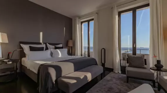 Un'elegante camera da letto con vista sul mare in pieno sole. Al centro della stanza si trova un grande letto con biancheria grigia e cuscini bianchi e grigio scuro. Ai lati del letto si trovano comodini con lampade da tavolo e una comoda poltrona accanto al letto.